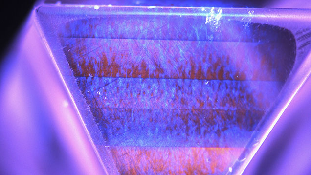 Figure 3. DiamondView image of one of the faces of the stone showing luminescence features consistent with a CVD synthetic diamond. Coarse bands of variable intensity orange emission from nitrogen vacancy centers are visible, along with growth striations and “plumes” of blue emission from dislocations within the bands. Image by Sema Firat; field of view 9.0 mm.