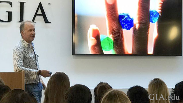 Brian Cook stands at a podium to the left of a big screen with an image of an upheld hand with brightly colored green and blue gems held between four fingers. Brian Cook stands at a podium to the left of a big screen with an image of an upheld hand with brightly colored green and blue gems held between four fingers.