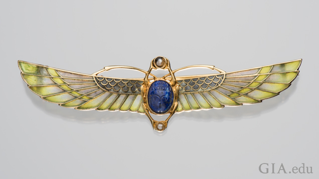 A scarab brooch with two sets of wings – one set is short, the other elongated. The body is a lapis lazuli and the long wings are a blue-green enamel. A scarab brooch with two sets of wings – one set is short, the other elongated. The body is a lapis lazuli and the long wings are a blue-green enamel.