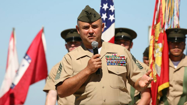 Manuel Diaz, in dress uniform, holds a microphone to address the crowd at his retirement ceremony. The American flag and U.S. Marine Corp flag are behind him, along with other flags and Marines. Manuel Diaz, in dress uniform, holds a microphone to address the crowd at his retirement ceremony. The American flag and U.S. Marine Corp flag are behind him, along with other flags and Marines.