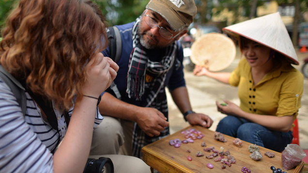 Manuel talks to two women as he looks at gems in a Vietnam gem market. The three sit around a small wooden table with gems separated into piles on it. Manuel talks to two women as he looks at gems in a Vietnam gem market. The three sit around a small wooden table with gems separated into piles on it.
