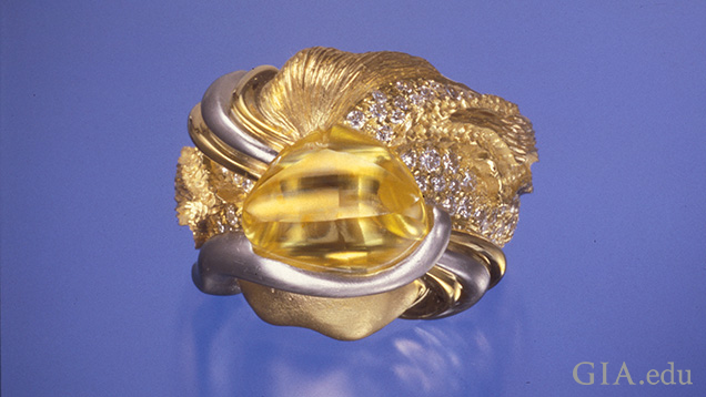 A polished, but not cut, triangular shaped yellow diamond sits in a nugget of gold that is diamond cut and set with diamonds and has ribbons of gold and silver running around it. A polished, but not cut, triangular shaped yellow diamond sits in a nugget of gold that is diamond cut and set with diamonds and has ribbons of gold and silver running around it.