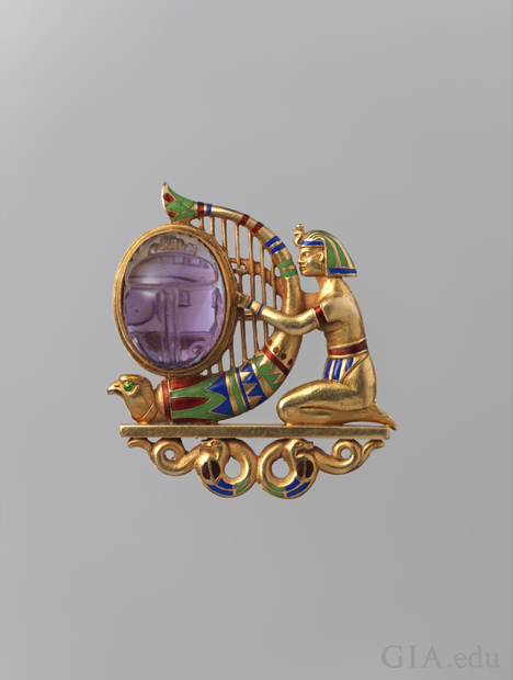 A kneeling figure clad in Egyptian headdress, arm bands, and loincloth playing a falcon-headed harp set with an oval amethyst carved as a scarab. The figure and harp rest on a plinth supported by two coiled snakes. Green, blue, and red enamel accent the h A kneeling figure clad in Egyptian headdress, arm bands, and loincloth playing a falcon-headed harp set with an oval amethyst carved as a scarab. The figure and harp rest on a plinth supported by two coiled snakes. Green, blue, and red enamel accent the h
