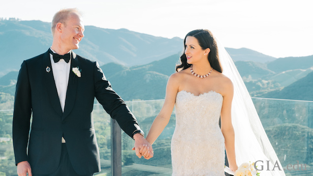 黒いタキシードを着た新郎と白いウェディングドレスを着た新婦が、結婚式で手を握り、見つめ合う。背景にはカリフォルニアの山々が見える。