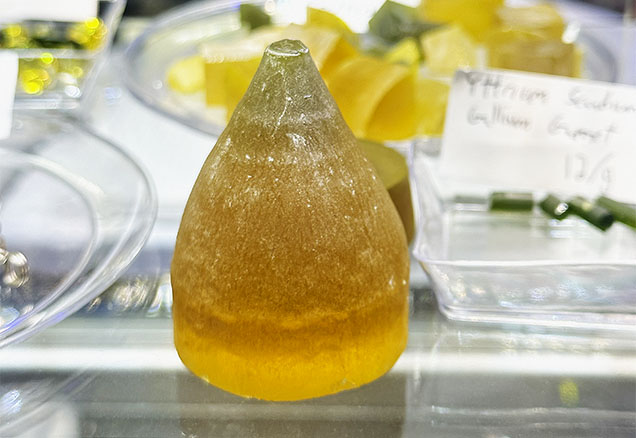Figure 2. The top of a YAG boule doped with cerium and neodymium and produced by the Czochralski method, measuring approximately 36 mm in diameter and 45 mm tall. Parts of the boule that cannot be used for scientific purposes can be sold to the gem and jewelry trade for faceting. Photo by Lisa Kennedy; courtesy of Turtle’s Hoard. Figure 2. The top of a YAG boule doped with cerium and neodymium and produced by the Czochralski method, measuring approximately 36 mm in diameter and 45 mm tall. Parts of the boule that cannot be used for scientific purposes can be sold to the gem and jewelry trade for faceting. Photo by Lisa Kennedy; courtesy of Turtle’s Hoard.