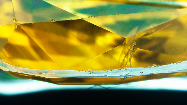 Figure 2. The appearance of laser ablation spots on a faceted yellow sapphire. There are two ablation pits on the girdle, located left and right of center. They are 55 μm in diameter and essentially undetectable without a microscope or loupe. Photomicrograph by Aaron Palke; field of view 5.21 mm. Figure 2. The appearance of laser ablation spots on a faceted yellow sapphire. There are two ablation pits on the girdle, located left and right of center. They are 55 μm in diameter and essentially undetectable without a microscope or loupe. Photomicrograph by Aaron Palke; field of view 5.21 mm.