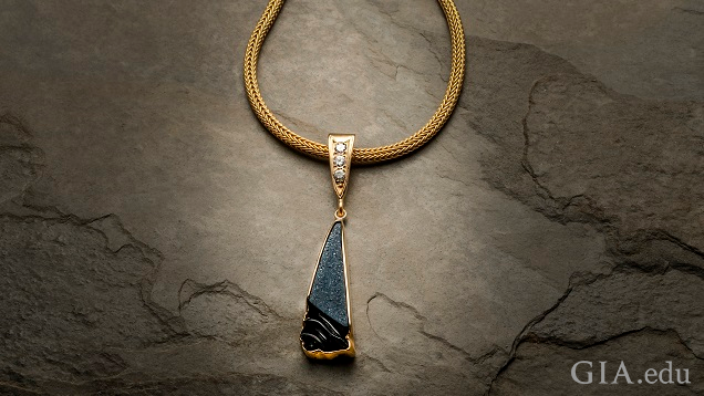 A triangle-shaped piece of black onyx drusy carving is framed in 14k yellow gold. It hangs from a triangle-shaped gold bail set with three 0.10 pt diamonds on a gold chain.