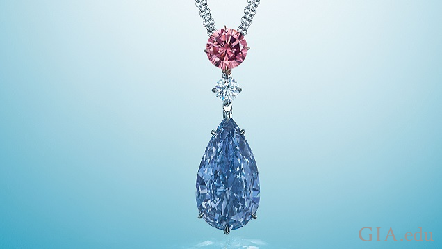 A large blue pear shaped diamond hangs from a colorless and pink diamond.