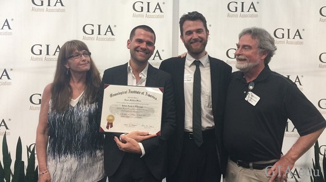 group of four people (mom, two sons and dad) stand in front of GIA backscreen with one son holding GIA diploma.