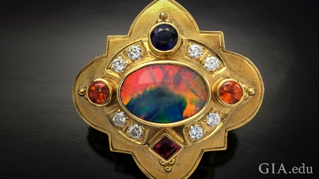 An orange, blue and green stone is the center piece of this shield-like brooch. The center stone is framed by diamonds, orange, blue and red gems. An orange, blue and green stone is the center piece of this shield-like brooch. The center stone is framed by diamonds, orange, blue and red gems.